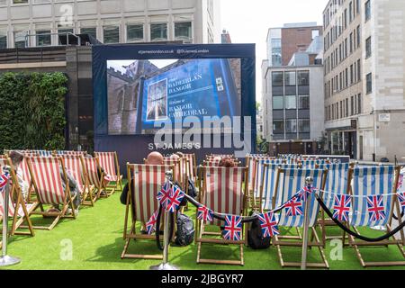 Liegestühle in einer Londoner Hinterstraße, mit Menschen, die sich hinsetzen und das Platinum Jubilee der Königin auf einem großen Bildschirm beobachten. London - 4.. Juni 2022 Stockfoto