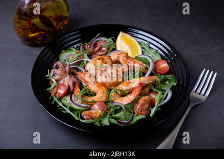 Salat mit Meeresfrüchten, Rucola, Tomaten, Gurken, roter Zwiebel und Zitrone auf einem schwarzen Teller. Schwarzer Hintergrund, Nahaufnahme. Stockfoto