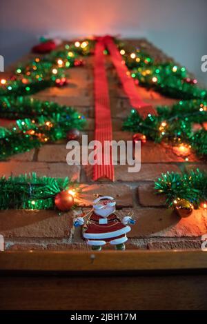 Weihnachtsdekoration über einem Holzofen mit bunten Lichtern und synthetischen Ästen, rotem Band über der Wand und einem kleinen Weihnachtsmann als ma Stockfoto