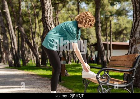 Schöne Rothaarige Frau in sportlicher Kleidung, die auf dem Stadtpark steht, im Freien Laufschuhe bindet, die auf der Parkbank gelehnt sind und sich zum Laufen vorbereiten. Joggi Stockfoto