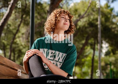 Nette kaukasische Frau trägt grünes T-Shirt im Stadtpark, Krämpfe im Bein im Freien und Schmerzen, berühren ihr Knie Knöchel schmerzhafte Verletzungen draußen Stockfoto