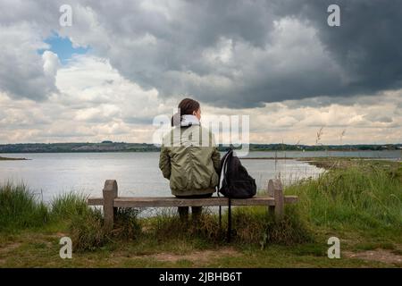 Rückansicht einer Frau, die allein auf einer Bank sitzt und den Blick über das Wasser und den stürmischen Himmel betrachtet. Stockfoto