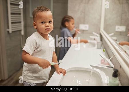 Porträt von zwei afroamerikanischen Kindern im Badezimmer konzentrieren sich auf kleine Kleinkind Jungen Zähneputzen und Blick auf Kamera, kopieren Raum Stockfoto