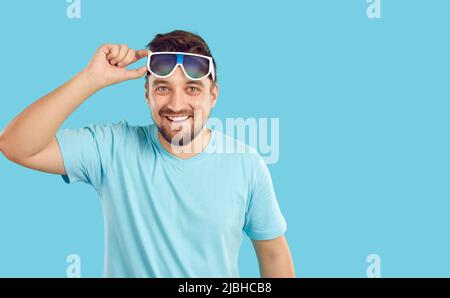 Ein glücklicher, gutaussehender Mann im blauen T-Shirt hebt seine Sonnenbrille, blickt auf die Kamera und lächelt Stockfoto