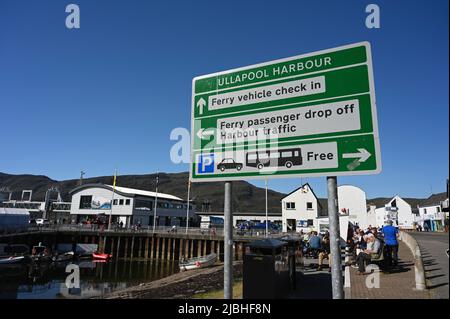 Schild für den Hafen Ullapool in Schottland, Großbritannien, mit Pfeilen zu den Fährgebieten. Menschen und Fährengebäude im Hintergrund. Blauer Himmel, sonniger Tag. Stockfoto