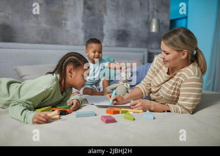 Porträt einer glücklichen kaukasischen Mutter mit zwei afroamerikanischen Kindern, die zusammen auf dem Bett spielen Stockfoto