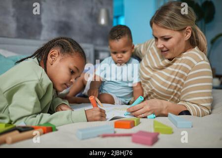 Porträt einer glücklichen kaukasischen Mutter mit zwei afroamerikanischen Kindern, die zusammen auf dem Bett liegen Stockfoto