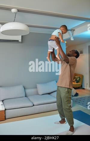In voller Länge Porträt von glücklichen schwarzen Vater spielen mit niedlichen Baby zu Hause und halten Sohn in der Luft Stockfoto