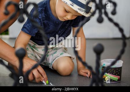 Der kleine Junge und seine Mutter zeichnen mit bunten Kreidekreiden auf einem betonierten Bürgersteig von oben. Die Mutter hilft, sich mit Hilfe ihrer Hand zu konzentrieren Stockfoto