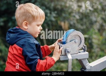 Der kleine Junge ist daran interessiert, mit der Boot-Anhängerwinde, der Bow-Rest, dem blauen Laderiemen und dessen Riemenscheibe und Zahnrädern zu spielen. Stockfoto