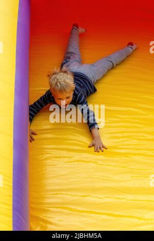 Ein mutiger kleiner Junge rutscht auf einem rot-gelben Trampolin in einem Vergnügungspark hinunter. Stockfoto