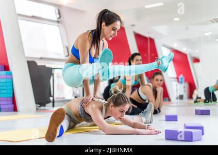 Zwei Mädchen, die Stretching-Übungen machen, arbeiten zu zweit mit einer Frau, die in einer weit nach vorne sitzenden, nach vorne gebeugeten Pose sitzt, und einer anderen Sportlerin, die Glühwürmchen macht Stockfoto