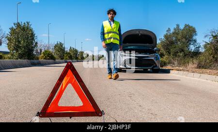 Junger Mann, der ein rotes Dreieck vorbereitet, um andere Verkehrsteilnehmer zu warnen, Autoausfall oder Motorausfall halten an der Landstraße an. Männlicher Fahrer, der in der Nähe eines b steht Stockfoto