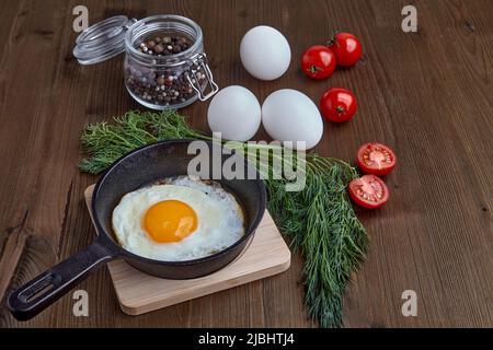 Gebratenes Hühnerei in einer kleinen Pfanne mit einem Ständer, einem Bund Dill, Kirschtomaten und ganzen weißen Eiern auf einem braunen Holztisch. Leichtes Frühstück Stockfoto