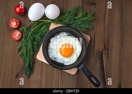 Ein gebratenes Hühnerei in einer kleinen Bratpfanne mit Ständer, ein Bund Dill, Kirschtomaten und ganze weiße Eier auf einem braunen Holztisch. Leichtes Frühstück Stockfoto