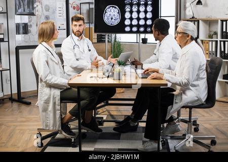 Ein Team internationaler medizinischer Experten sitzt am Tisch und analysiert den MRT-Scan des Patienten auf dem Fernsehbildschirm. Gruppe von multiethnischen männlichen und weiblichen Ärzten mit modernen Gadgets während der Arbeit. Stockfoto