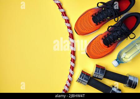 Trainingsgeräte. Orangefarbene Sport-Sneakers, Turnschuh in ein Maßband mit einem Meter eingewickelt, Hanteln und eine Flasche Wasser auf einem gelben Rücken Stockfoto