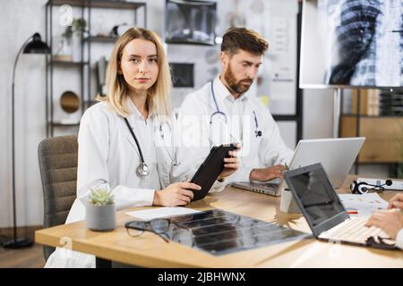 Porträt einer kaukasischen Ärztin, die während eines Treffens des medizinischen Personals mit einem digitalen Tablet in den Händen am Tisch sitzt. Bärtig konzentrierter männlicher Arzt, der auf dem Hintergrund am Laptop arbeitet. Stockfoto