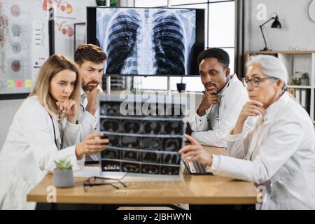 Nachdenkliche multirassische Ärzte untersuchen Patienten CT-Scan von Brust- und Bauchhöhle, während sie um den Schreibtisch sitzen. Pulmonologie, Lungenpathologie und Tuberk Stockfoto