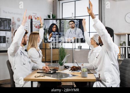 Team multiethnischer Ärzte im Gesundheitswesen, die während eines Online-Videotreffens mit erhobenen Armen am Tisch sitzen. Medizinische Mitarbeiter in weißen Laborkittel stimmen für ein neues erfolgreiches tomographisches Projekt in einer modernen Klinik. Stockfoto