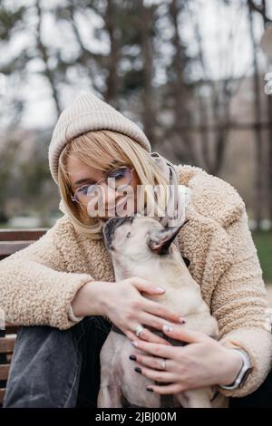 Hübsche Blondine umarmt ihren niedlichen Mops. Schöne junge Frau, die draußen mit ihrem Haustier in den Händen sitzt. Stockfoto