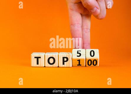 Von Top 100 bis 50 Symbol. Geschäftsmann dreht Holzwürfel und ändert die Begriffe Top 100 zu Top 50. Schöner orangefarbener Tisch und Hintergrund. Unternehmen Stockfoto