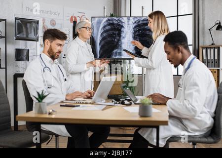 Eine Gruppe von zwei Ärzten im unterschiedlichen Alter, die über Röntgenergebnisse sprechen, während multiethnische Männer an Laptops arbeiten. Kompetente Mitarbeiter geben Ratschläge und teilen Wissen im modernen Sitzungssaal der Klinik. Stockfoto