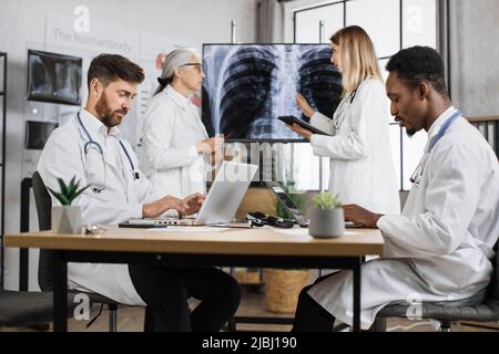 Team aus zwei mehrethnischen männlichen medizinischen Mitarbeitern, die auf drahtlosen Laptops tippen, während weibliche Kollegen Tomographieergebnisse diskutieren und Diagnosen stellen. H Stockfoto