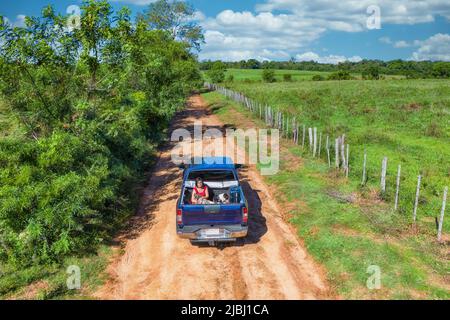 Luftaufnahme einer Frau, die mit ihrem Hund und ihrem Koffer auf einer typischen roten Sandstraße in Paraguay auf der Rückseite eines Pickup-Trucks sitzt. Stockfoto