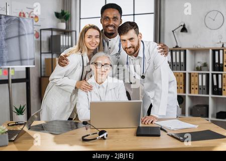 Eine Gruppe multikultureller medizinischer Mitarbeiter in weißen Laborkittel umarmt und blickt mit einem Lächeln in den hellen Sitzungssaal der Klinik auf die Kamera. Effektives Teamwork und Konzept der Zusammenarbeit im Gesundheitswesen. Stockfoto
