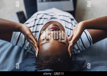 Nahaufnahme einer Frau, die den Kopf greift und einen Laptop benutzt, während sie auf dem Boden sitzt. Schöne schwarze Frau. Nahaufnahme, Draufsicht. Wunderschönes afrikanisch Stockfoto