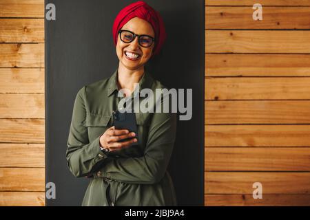 Glücklicher muslimischer Unternehmer, der die Kamera anlächelt, während er ein Smartphone hält. Fröhliche Geschäftsfrau beim Lesen einer Textnachricht in einem Büro. Weibliche Entrepr Stockfoto