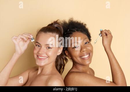 Zwei wunderschöne kaukasische und afroamerikanische Frauen halten Pipetten mit Serum mit Vitamin C und Hyaluronsäure. Feuchtigkeitsspendende und Hautpflege. Stockfoto