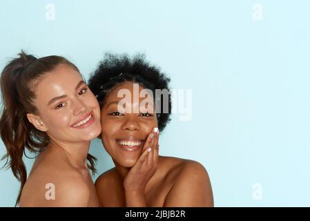 Porträt zweier junger Frauen mit einer perfekten feuchtigkeitsspendenden Haut, die zusammen stehen und vor blauem Hintergrund die Kamera anlächeln. Die Konz Stockfoto