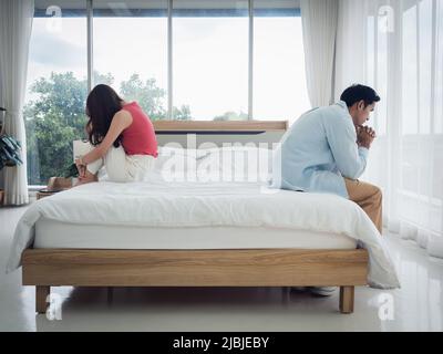 Die Angst der asiatischen Paarliebhaber auf dem Bett. Nachdenklicher Mann und junge Frau sitzen auf dem Bett mit Beziehungsschwierigkeiten und fühlen sich traurig und thinki Stockfoto