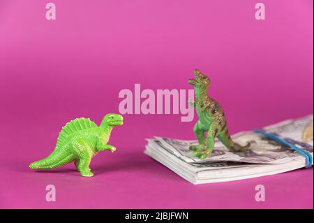 Dinosaurier-Miniaturen und ein Wad von hundert-Dollar-Scheinen vor purpurem Hintergrund. Ein Geldstapel, der mit einem Gummiband gebunden ist. Grüne Figuren von Tieren Stockfoto
