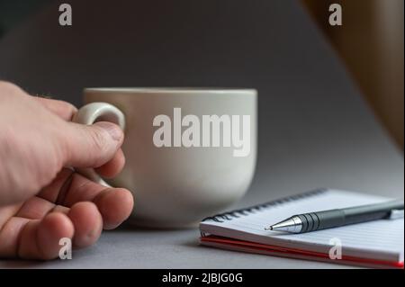 Nahaufnahme der Hand eines Mannes mit einem beigefarbenen Keramikbecher und einem Griff. Öffnen Sie das federbelastete Notizbuch und den Stift nebeneinander. Seitenansicht. Selektiver Fokus Stockfoto