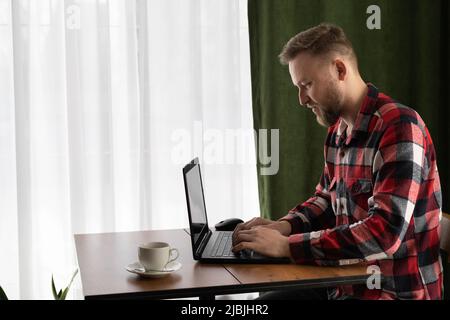 Schöner junger Mann, Freiberufler, der Laptop benutzt, von zu Hause aus arbeitet, online studiert, Typ, der am pc-Laptop tippt, im Internet surft, auf den Bildschirm schaut und Spaß hat Stockfoto