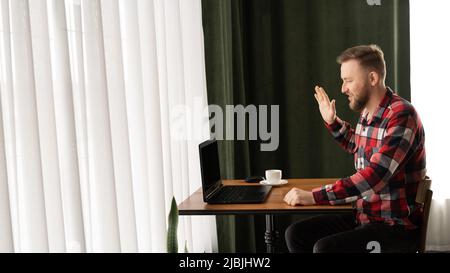 Glücklicher Tausendjähriger Kaukasianer in legerer Kleidung, der einen Videoanruf macht, während er zu Hause mit einem Laptop an einem Tisch sitzt. Hallo. mit erhobener Hand winkend Banner Stockfoto