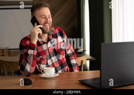 Kaukasischer lächelnder Mann im roten Hemd mit Laptop zum Online-Shopping, zum Telefonieren und Kaffeetrinken während der Arbeit als freier Online-Mitarbeiter Stockfoto