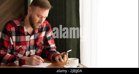 Digitales Lernen, Online-Studium, Gelegenheitlicher Mann oder Student, der am Smartphone arbeitet und auf einem Notizblock auf Papier schreibt, junger Mann, der im Café oder zu Hause ein Mobiltelefon benutzt Stockfoto