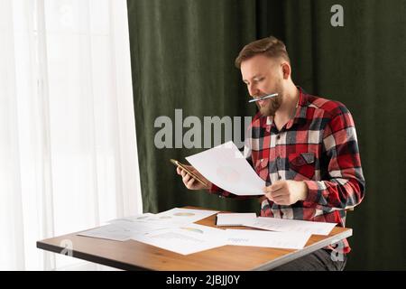 Konzentrierter kaukasischer Mann in Freizeitkleidung, der sich durch den Marktforschungsbericht durchschaut, statistische Daten in Diagrammen analysiert und eine Wachstumsstrategie entwickelt Stockfoto