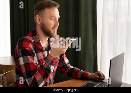 Mann, der im Büro sitzt und auf einem Freisprechtelefon spricht, Spracherkennung macht, Internetdienste über einen virtuellen Assistenten nutzt, Audionachrichten aufnimmt Stockfoto