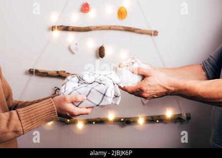 Frau und Mann geben furoshiki Verpackung Geschenkboxen einander gegen Dekoration Weihnachtsbaum aus trockenen Stäbchen, trocken orange, Zapfen auf weißem Rücken Stockfoto