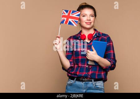 Attraktive Studentin hält kleine britische Flagge und Notizbuch im Studio auf beigem Hintergrund. Studienkonzept im Ausland. Internationales Austauschprogramm für Studenten. Stockfoto