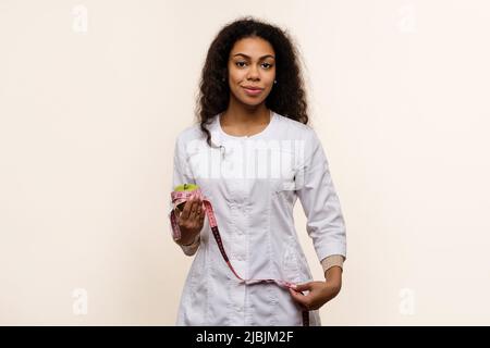 Afroamerikanische Ernährungswissenschaftlerin hält grünen Apfel und Messband auf hellem Hintergrund. Friendly Dark skinned in Uniform fördert das Konzept von He Stockfoto