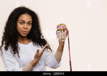 Nahaufnahme Porträt afroamerikanische Ernährungswissenschaftlerin auf hellem Hintergrund im Studio. Der Arzt zeigt auf einen grünen Apfel, der in einem Maßband in der Hand eingewickelt ist. Stockfoto