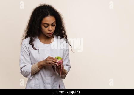 Dunkelhäutig Ernährungsberaterin in Uniform sieht grüner Apfel und Messband in den Händen aus. Stockfoto