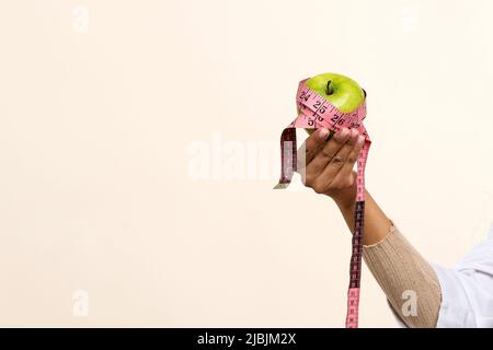 Konzept der richtigen Ernährung und einer gesunden Lebensweise. Grüner Apfel, in rosa Maßband eingewickelt, in der Hand von dunkelhäutiger, nicht erkennbarer, gesichtsloser Nutritio Stockfoto