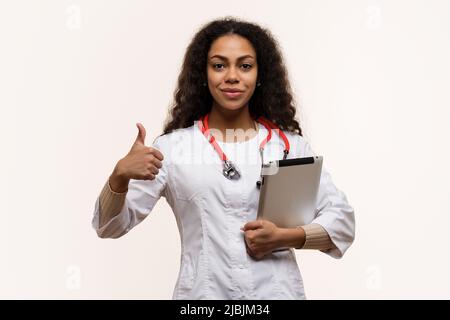 Dunkelhäutig Arzt in Uniform mit Stethoskop zeigt Daumen nach oben wie auf hellem Hintergrund. African American Female General Doctor mit Tablet-PC. Dat Stockfoto